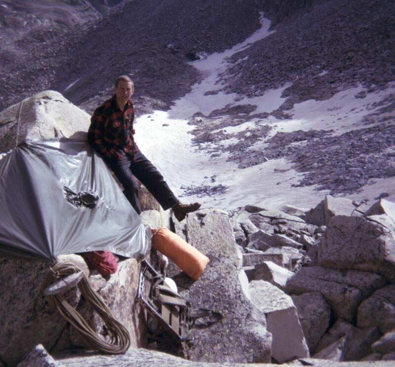 A man in a red and black flannel shirt and dark pants sits on a rock next to a makeshift shelter covered in a gray tarp. Climbing ropes and gear are visible near the shelter. The background shows a rocky, mountainous landscape with patches of snow.